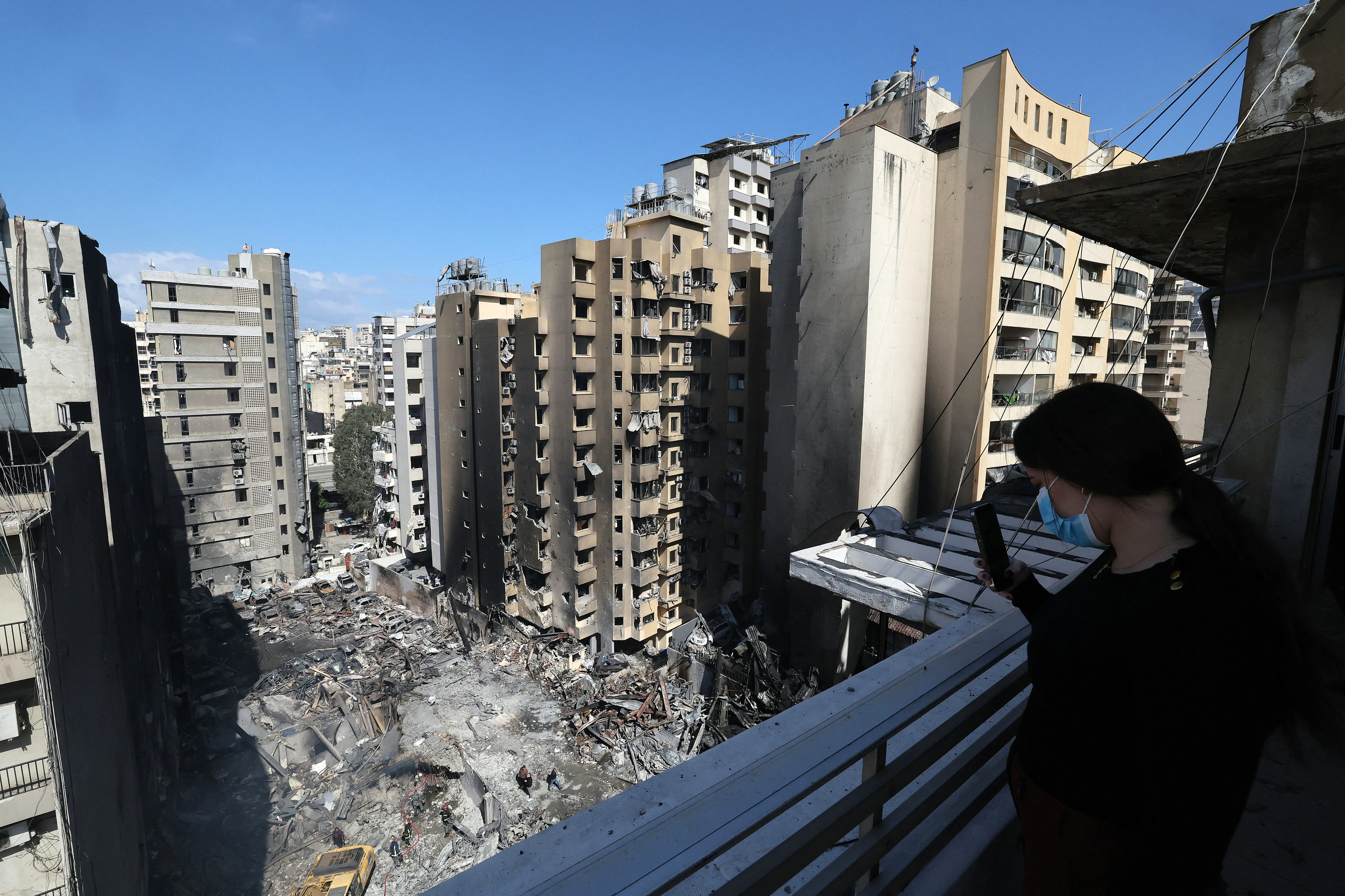 A woman wearing a face mask on a balcony takes a picture with her phone of Lebanese first responders searching under the rubble