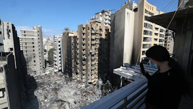 A woman wearing a face mask on a balcony takes a picture with her phone of Lebanese first responders searching under the rubble