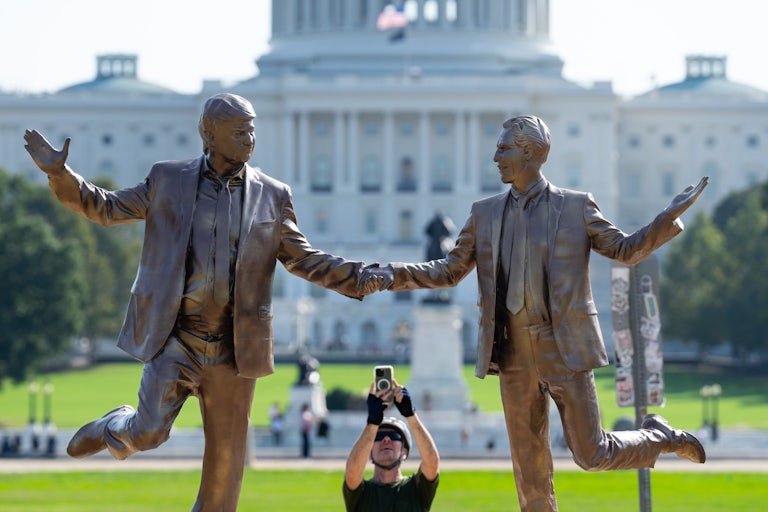 Statue of Donald Trump and Jeffrey Epstein frolicking and holding hands on the National Mall. A man takes a photo on his phone.