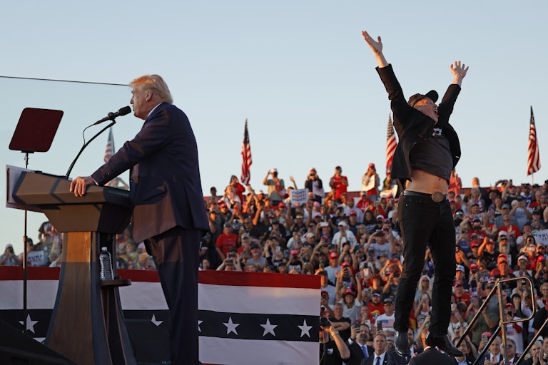 Donald Trump speaks at a podium during a campaign rally while Elon Musk jumps behind him