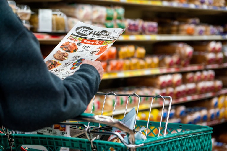 A shopper holds the weekly sales advertisement while pushing their cart in the grocery store.