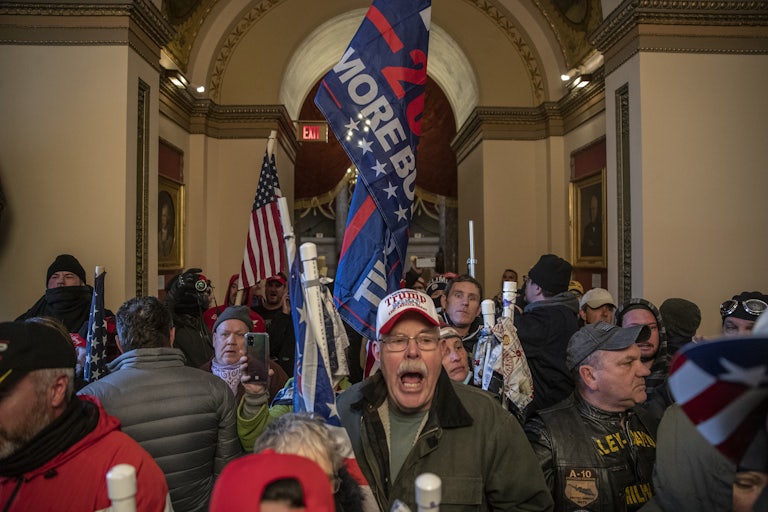 An old white man in the Capitol on January 6, 2021 wears a Trump cap and yells. Others around him hold U.S. flags and Trump 2020 flags.