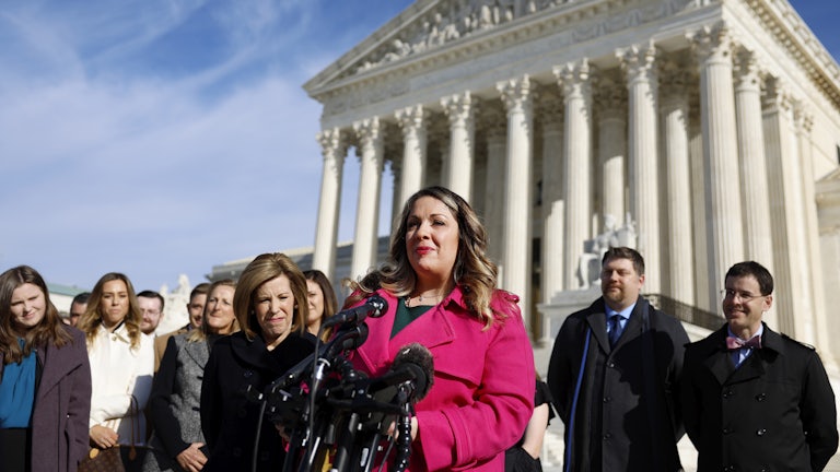 A woman in a pink coat stands at a podium in front of the Supreme Court building, flanked by supporters.