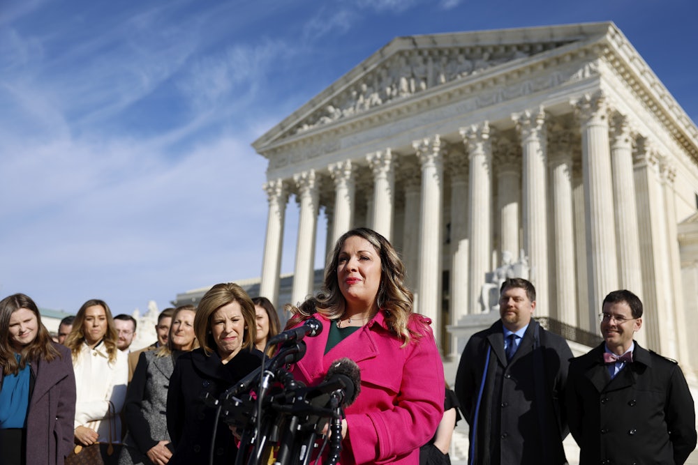A woman in a pink coat stands at a podium in front of the Supreme Court building, flanked by supporters.