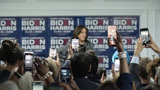 Kamala Harris stands at the front of a room facing a sea of people holding up cell phones to take pictures.