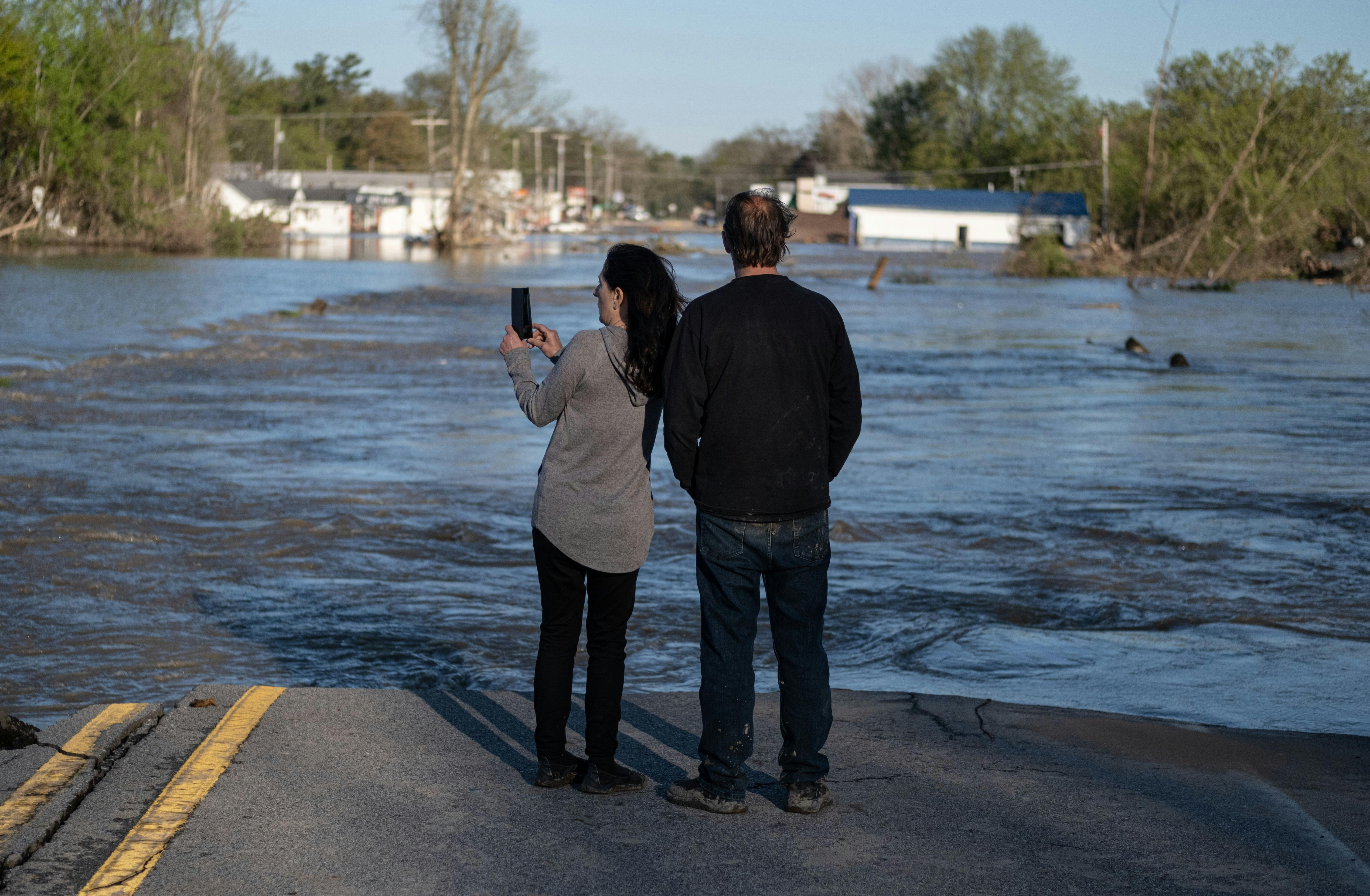 Two people stand at the edge of water flowing over a road, with flooded houses in the background.