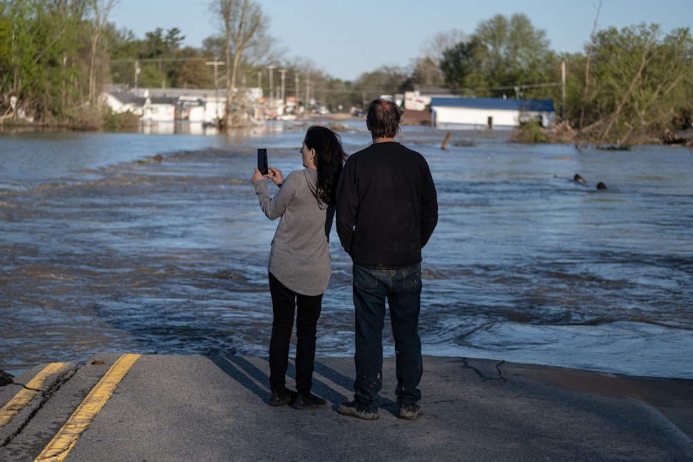 Two people stand at the edge of water flowing over a road, with flooded houses in the background.