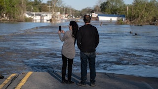 Two people stand at the edge of water flowing over a road, with flooded houses in the background.