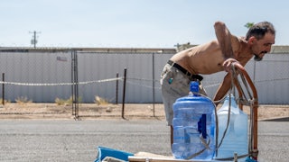 A person with no shirt rests their hands on their knees in the middle of a bright street while pushing a cart with a jug of water on top.