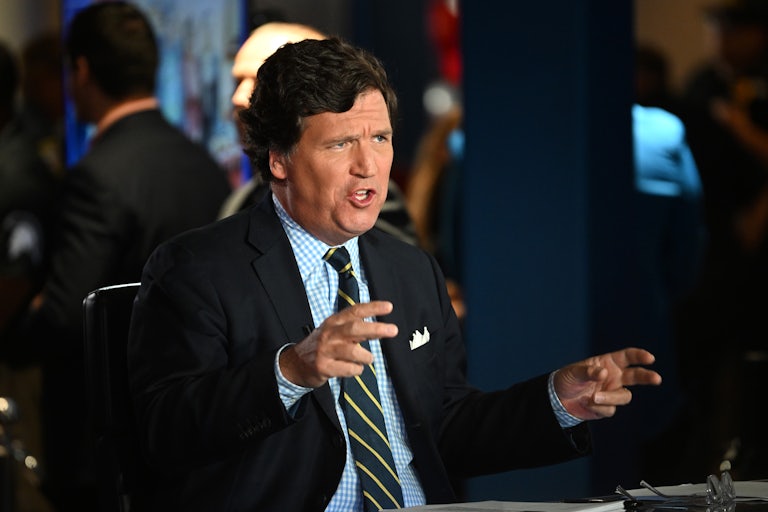 Tucker Carlson sitting at a table makes a weird face and air quotes with both hands