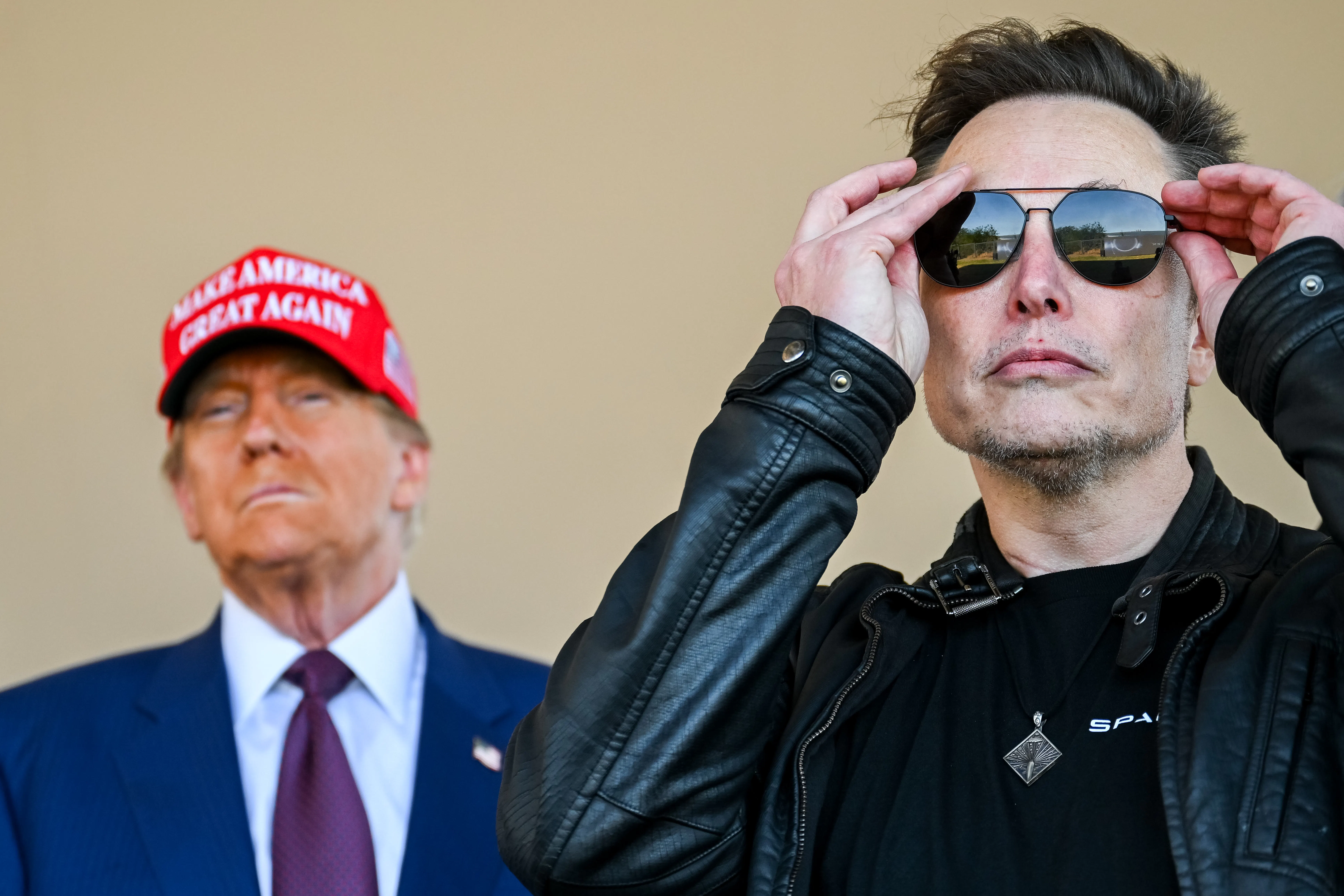 Musk puts on sunglasses while Trump stands behind him