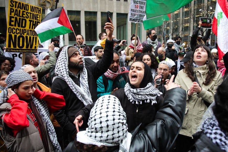 Protesters gather. Many wear the keffiyeh, wave Palestinian flag, or hold up signs that read "Stand with Palestine! End the Occupation Now!" One woman in the forefront wears a hijab and keffiyeh and yells.
