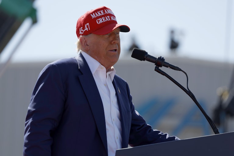 Donald Trump wears a MAGA hat and speaks at a lectern outdoors