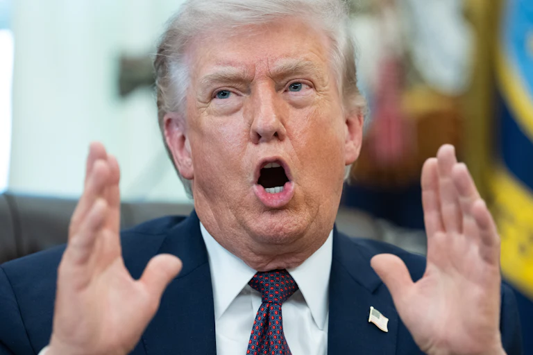 Donald Trump holds both hands next to his face and speaks while sitting at his desk in the Oval Office