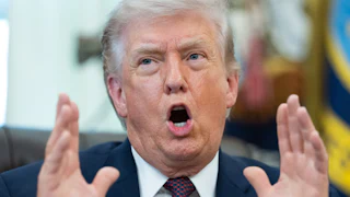 Donald Trump holds both hands next to his face and speaks while sitting at his desk in the Oval Office