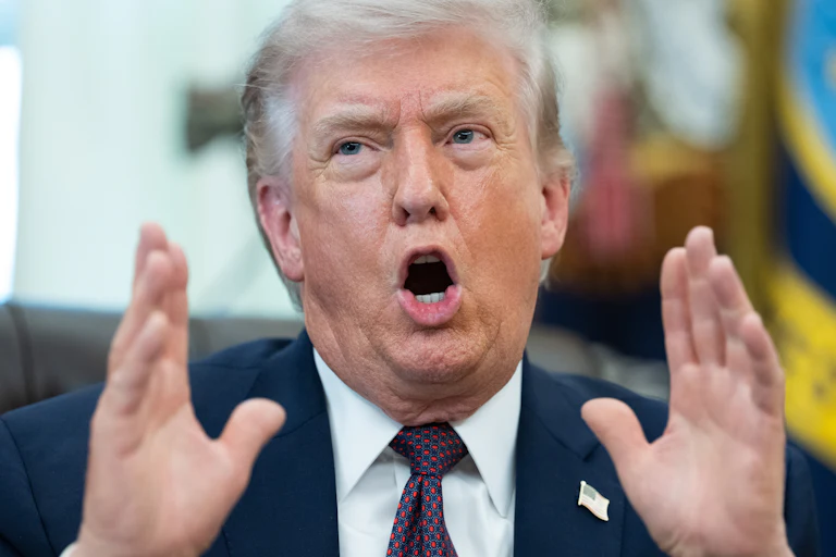 Donald Trump holds both hands next to his face and speaks while sitting at his desk in the Oval Office