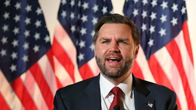 Vice President JD Vance speaks into a microphone while standing in front of American flags