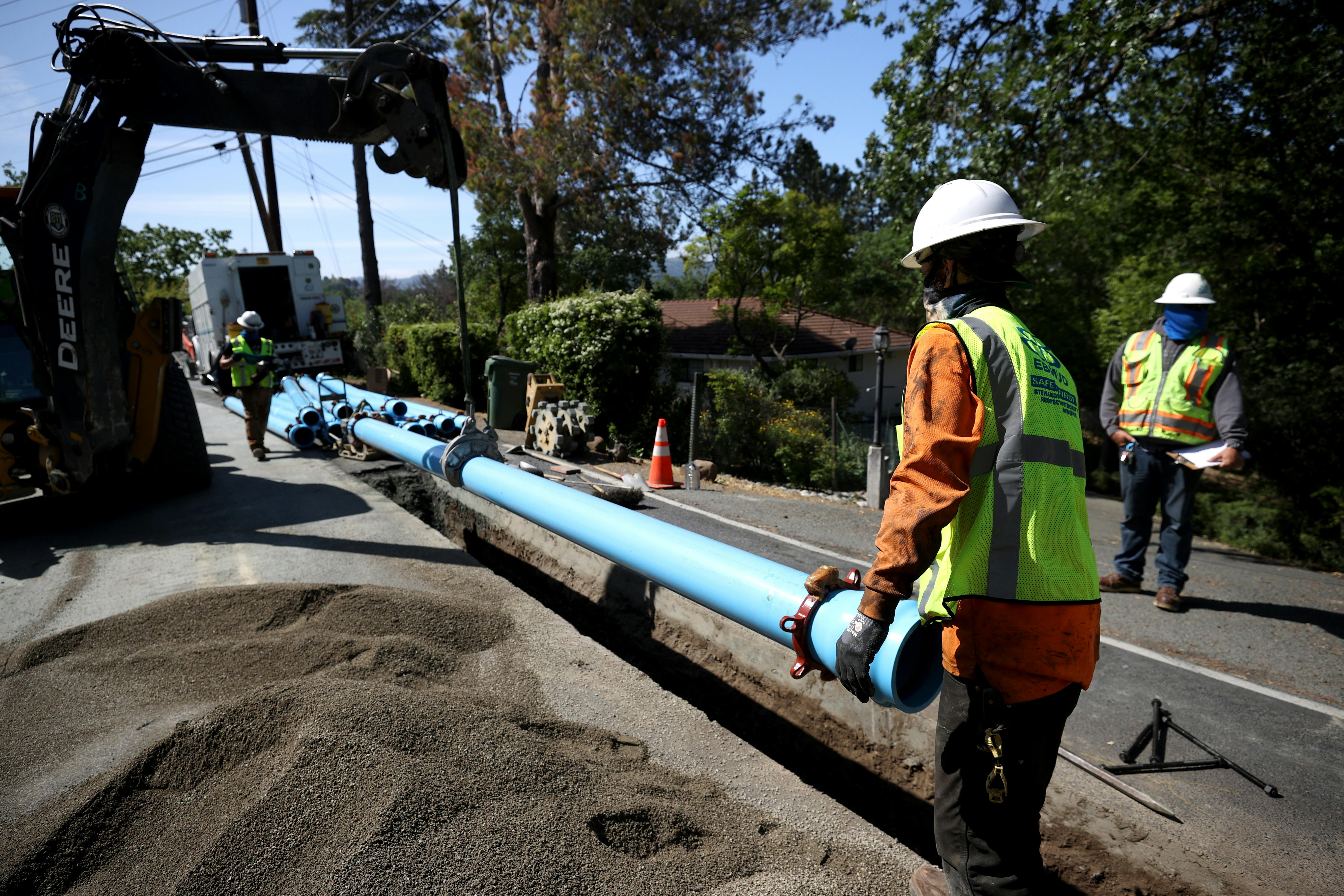A worker in a hard hat helps lower a pipe into a trench that has been dug in the middle of a road.