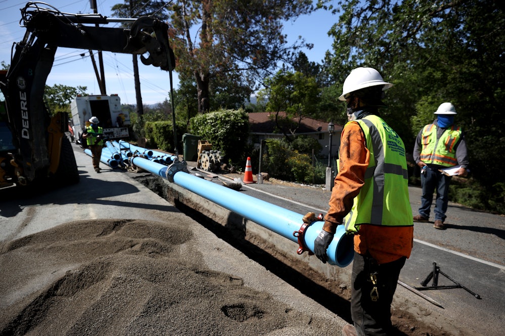 A worker in a hard hat helps lower a pipe into a trench that has been dug in the middle of a road.