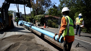 A worker in a hard hat helps lower a pipe into a trench that has been dug in the middle of a road.