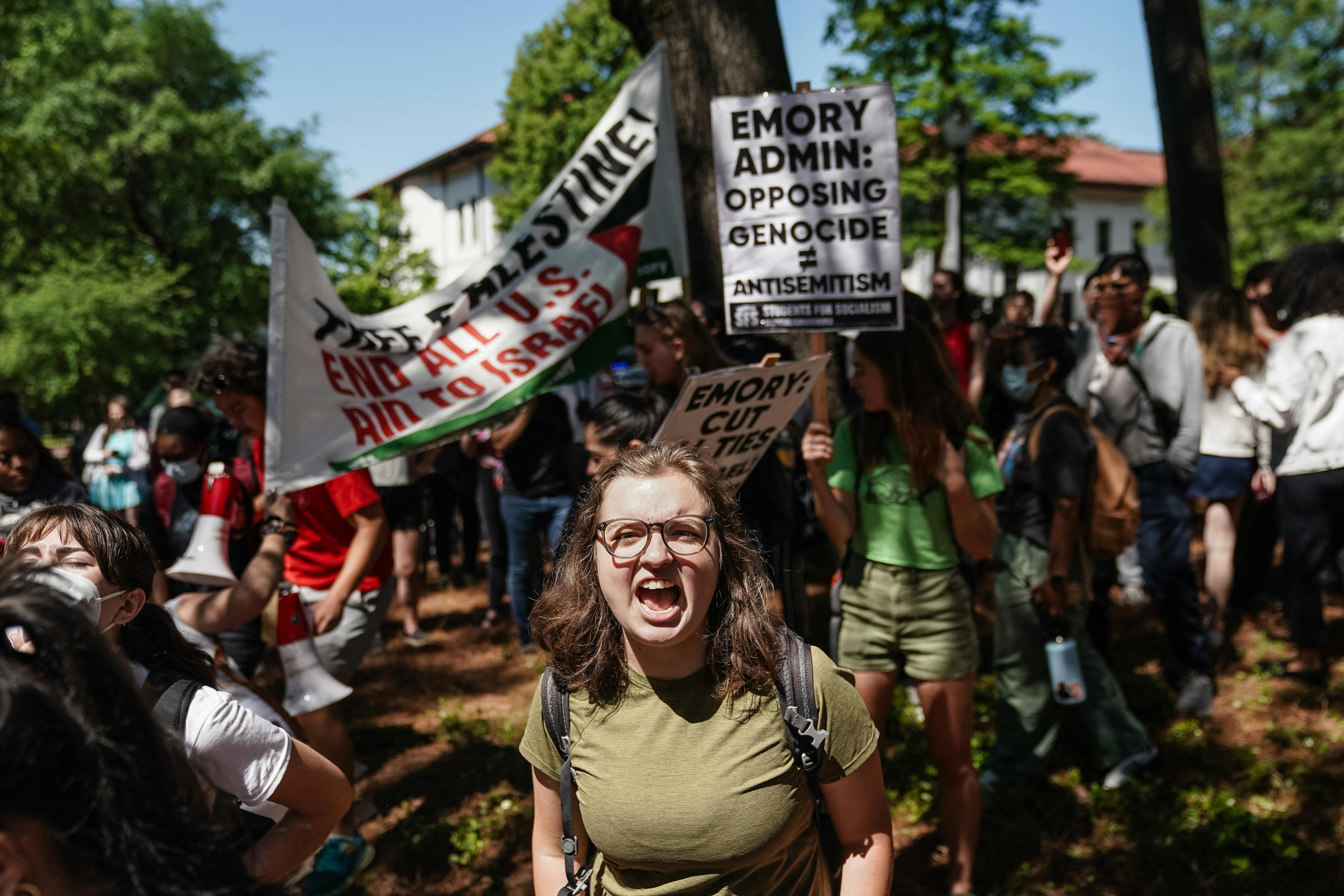 A young woman yells as she stands in front of several demonstrators holding banners that read "Emory admin: Opposing genocide [does not equal] antisemitism" and "End all U.S. aid to Israel." 