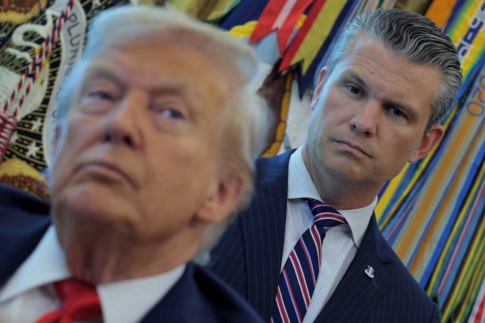 Defense Secretary Pete Hegseth looms behing President Donald Trump as he talks to journalists after signing executive orders in the Oval Office.