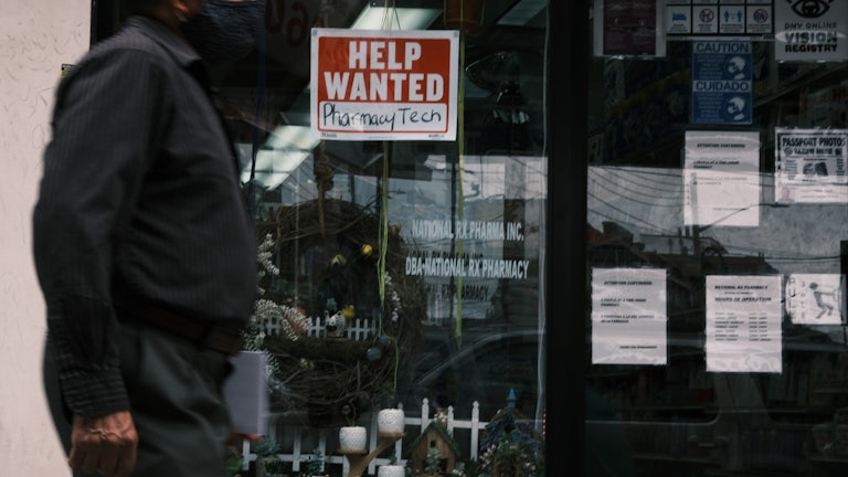 A masked man walks past a help wanted sign on a storefront window.