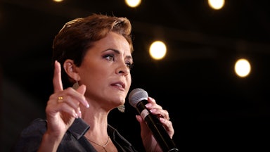 Kari Lake speaks at a campaign rally. She holds a mic in her left hand and is pointing the index finger on her right.