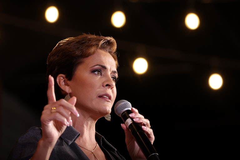 Kari Lake speaks at a campaign rally. She holds a mic in her left hand and is pointing the index finger on her right.