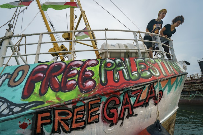 Two people on board the Freedom Aid Flotilla look down over the railing. The ship has graffiti that reads "Free Palestine" and "Free Gaza."