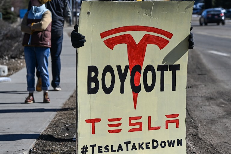 Someone holds a large sign that reads "Boycott Tesla #TeslaTakeDown."