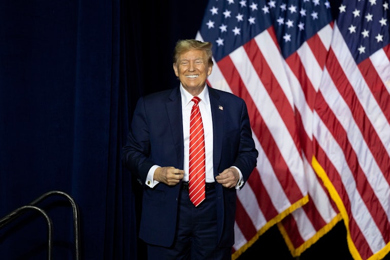 Donald Trump smiles and stands before some U.S. flags.