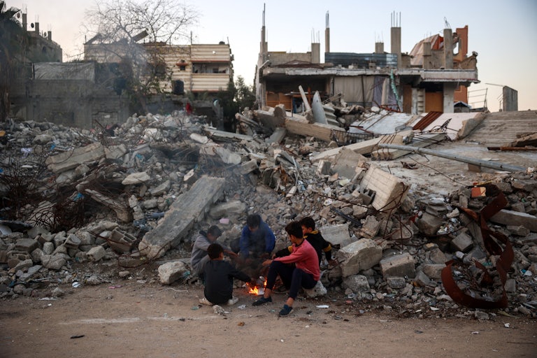 A group of children sit by a fire amid a larger scene of rubble