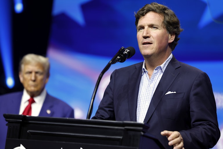 Tucker Carlson delivers a speech at a lectern. Donald Trump can be seen in the background.
