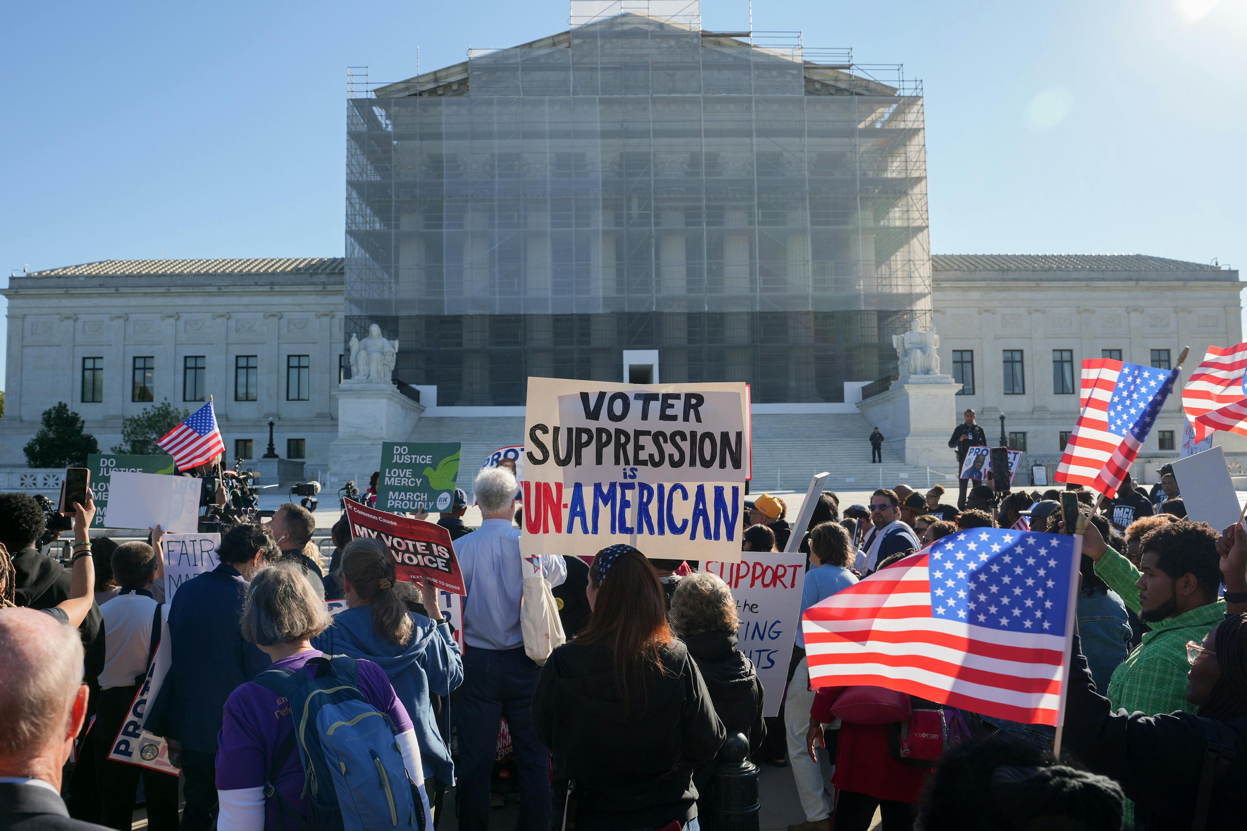 Protesters hold up U.S. flags and a sign that reads "Voter Suppression is Unamerican" while standing in front of the Supreme Court.
