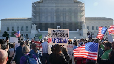 Protesters hold up U.S. flags and a sign that reads "Voter Suppression is Unamerican" while standing in front of the Supreme Court.