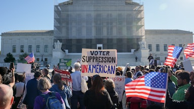 Protesters hold up U.S. flags and a sign that reads "Voter Suppression is Unamerican" while standing in front of the Supreme Court.
