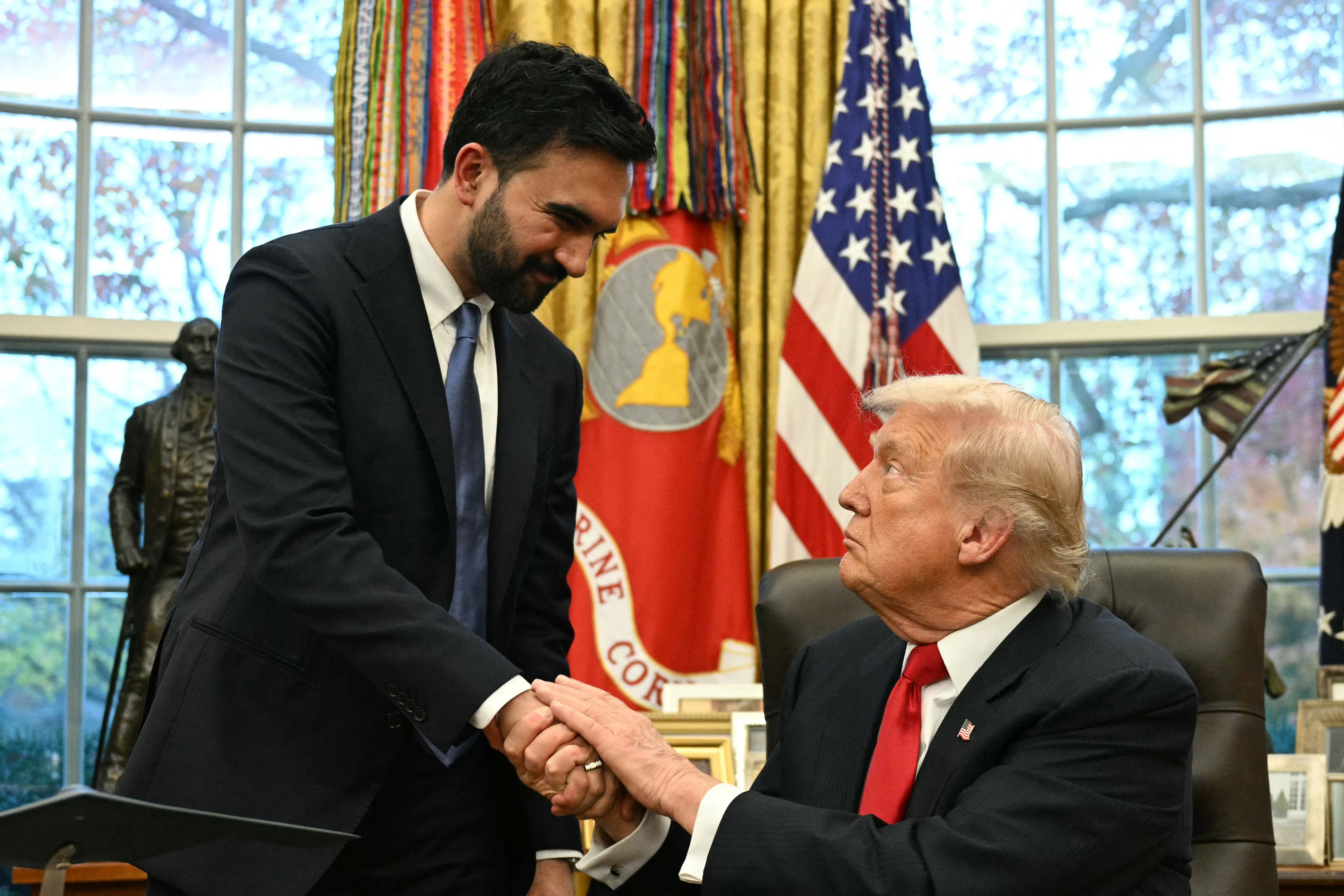 Zohran Mamdani, wearing a suit and tie, standing and shaking hands with a seated Donald Trump at his desk in the Oval Office, wearing a dark suit and red tie. Trump has a serious expression on his face.