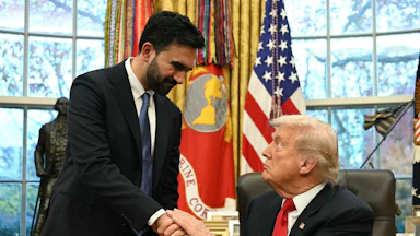 Zohran Mamdani, wearing a suit and tie, standing and shaking hands with a seated Donald Trump at his desk in the Oval Office, wearing a dark suit and red tie. Trump has a serious expression on his face.