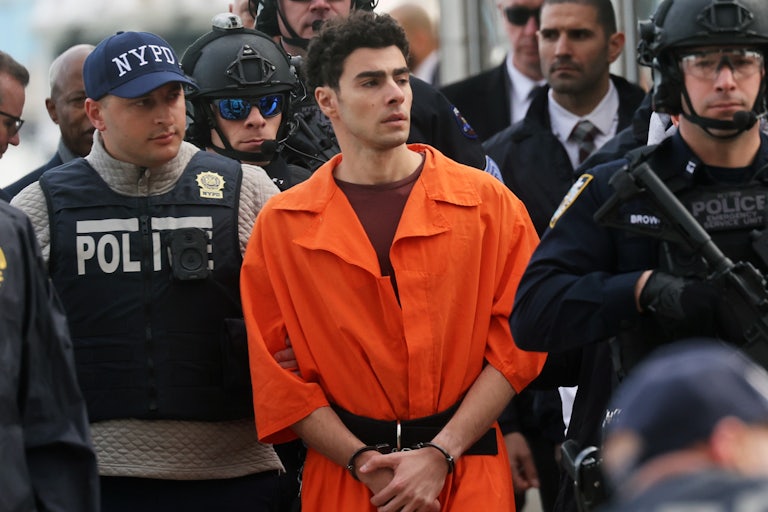 Luigi Mangione in an orange jumpsuit being escorted by law enforcement officials. (A corner of Eric Adams’s face can be seen.)