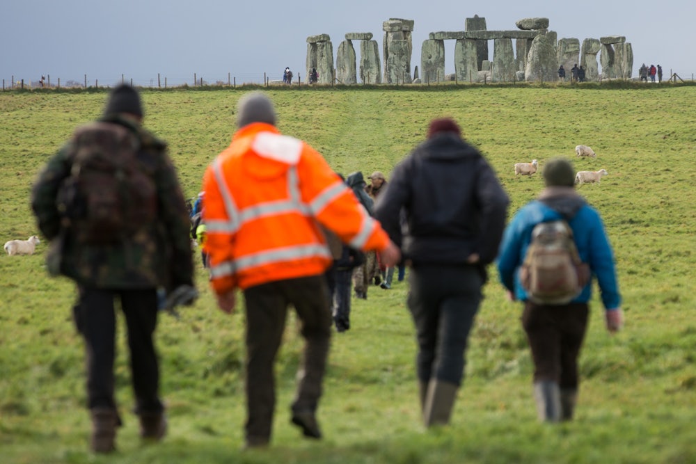 People wearing jackets walk across the field to Stonehenge, which stands in the background.