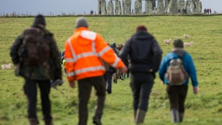 People wearing jackets walk across the field to Stonehenge, which stands in the background.