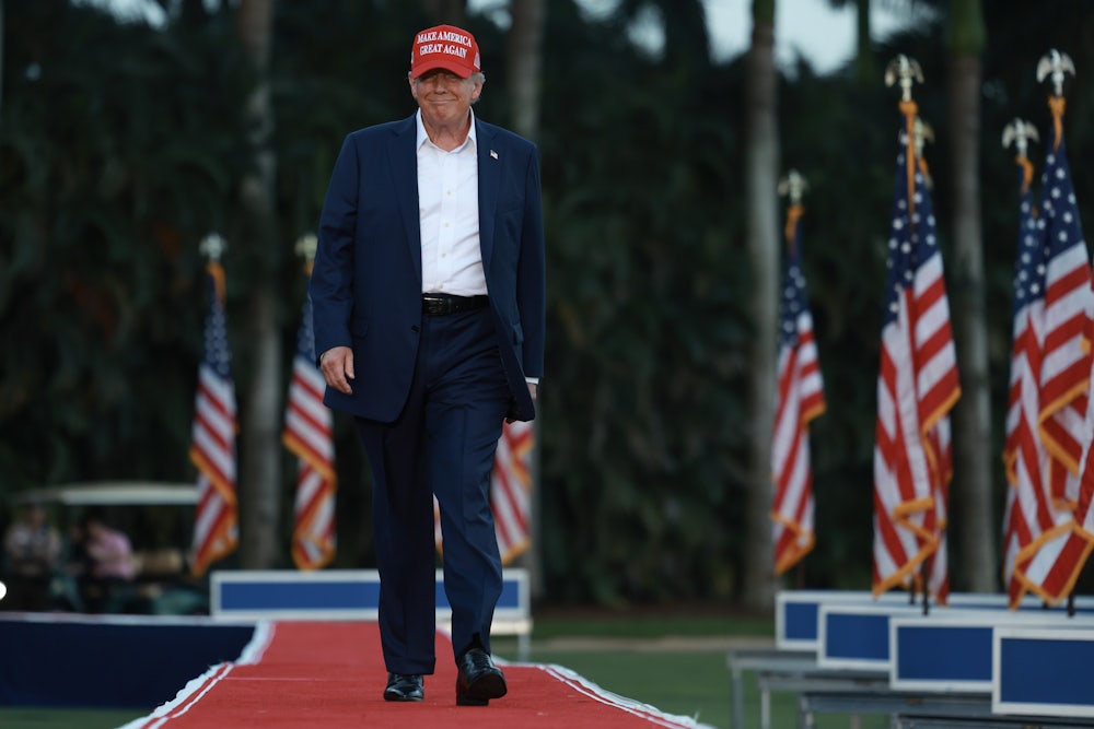 Trump walks across an elevated walkway, smiling while wearing a Make America Great Again hat.