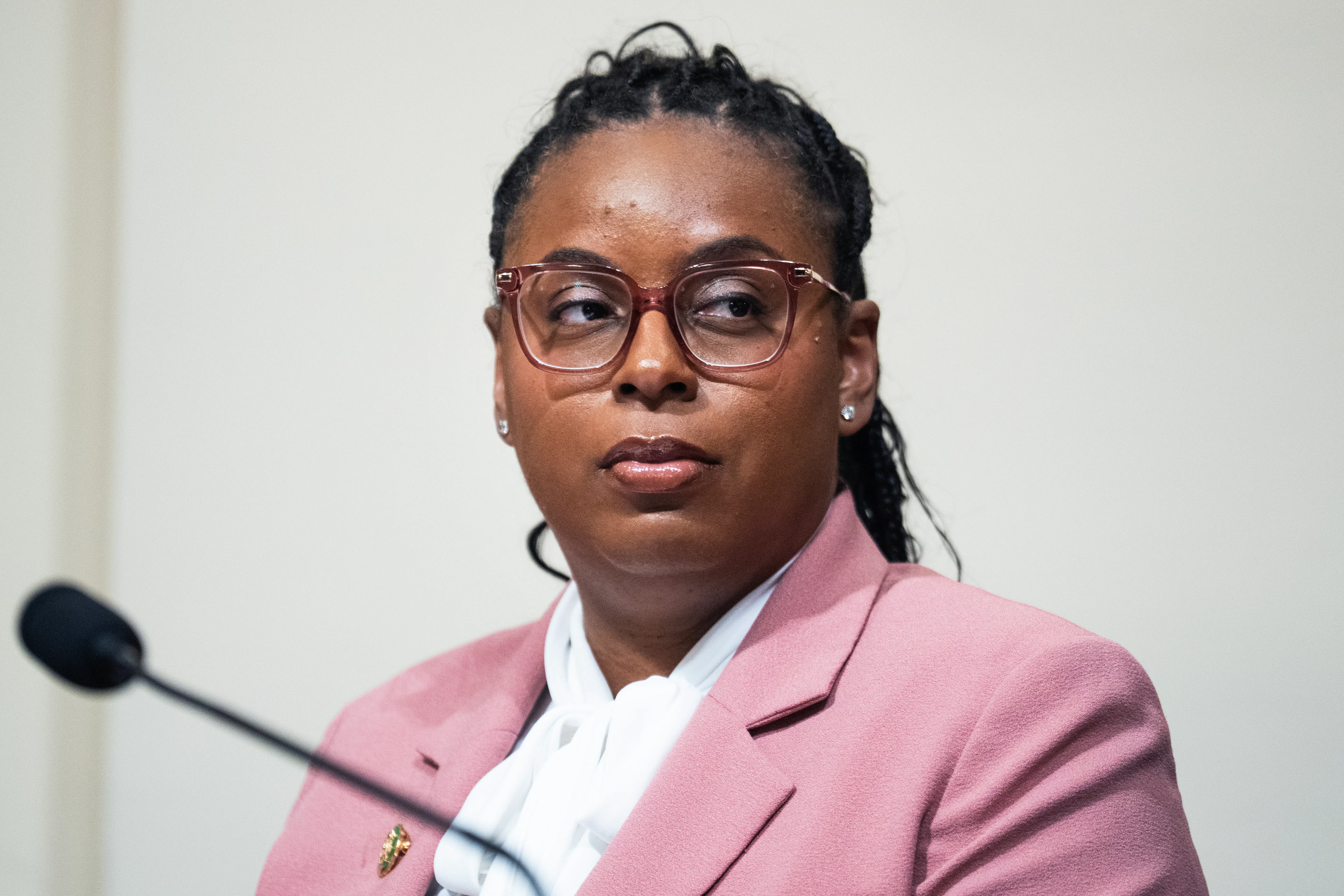 LaMonica McIver is seated, wearing a beige blazer and a white shirt with a microphone in front of her. 