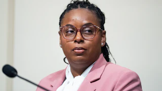 LaMonica McIver is seated, wearing a beige blazer and a white shirt with a microphone in front of her.