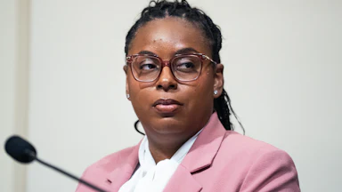 LaMonica McIver is seated, wearing a beige blazer and a white shirt with a microphone in front of her.