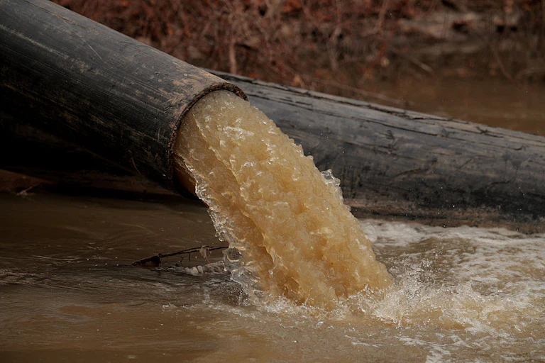 Brown water pours out of a pipe, against a riverbank.
