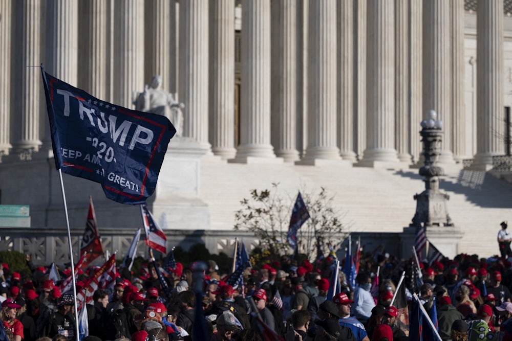 Many people gather before the steps and columns of the Supreme Court building, one person holding a large Trump banner.