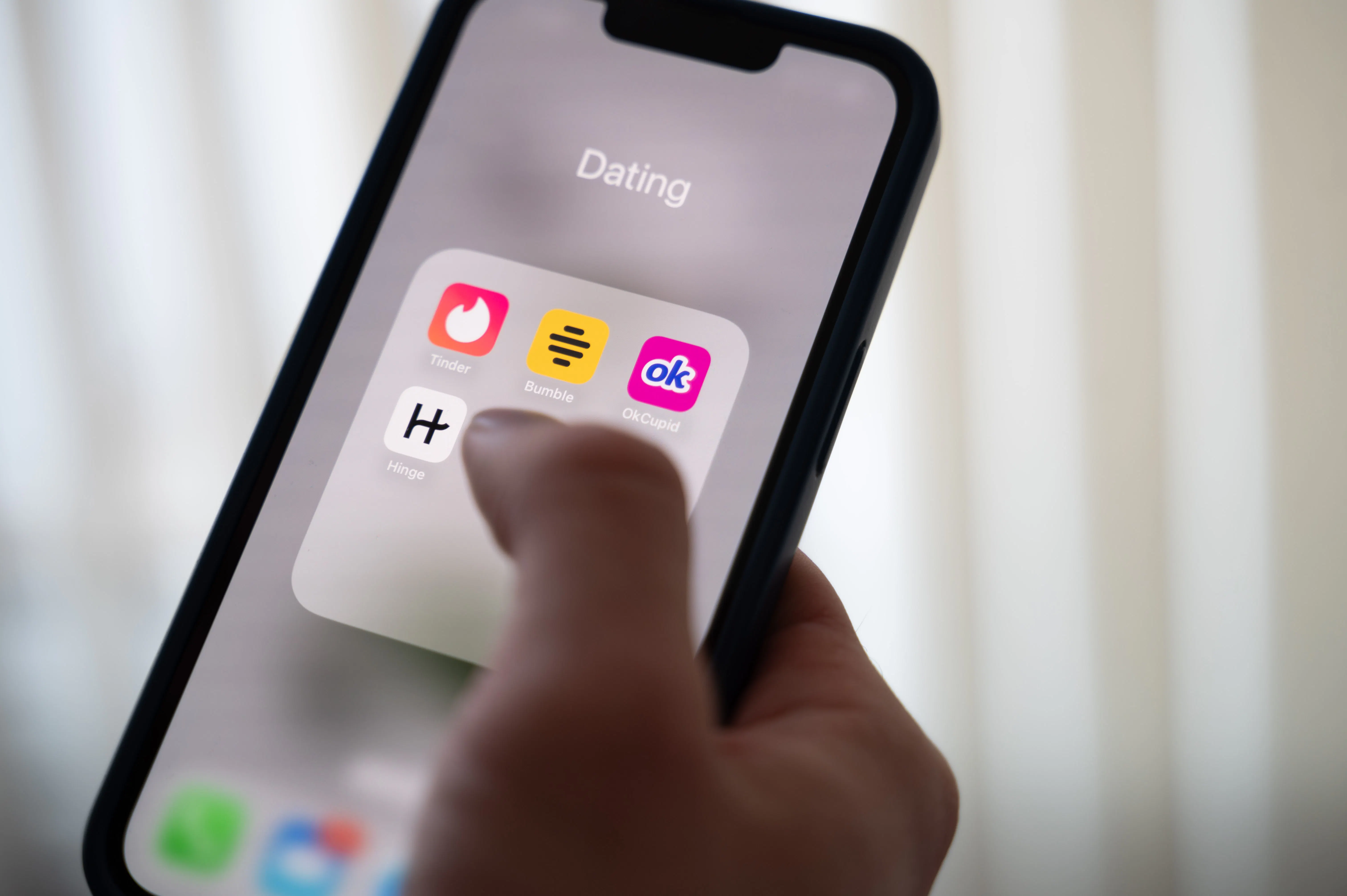 A hand holds a phone with several dating app son the screen: Tinder, Bumble, OkCupid, and Hinge.