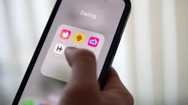 A hand holds a phone with several dating app son the screen: Tinder, Bumble, OkCupid, and Hinge.